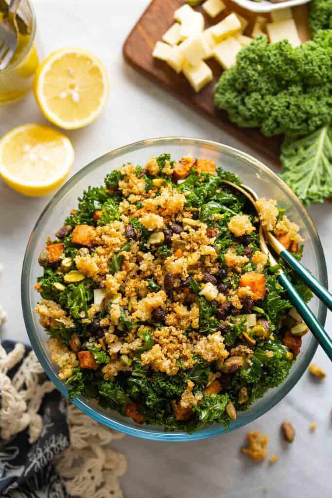 A salad made with roasted sweet potato & black beans, kale, cheese and chopped pistachios lot crispy parmesan quinoa in a glass salad bowl with serving spoons surrounded by the raw ingredients.