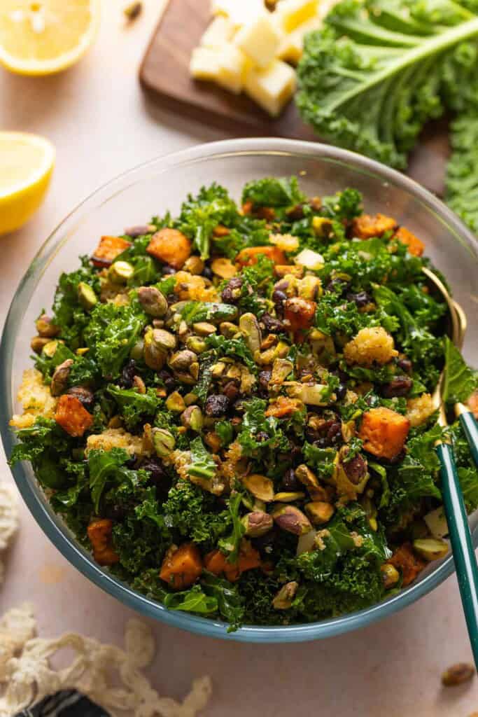 A salad made with roasted sweet potato & black beans, crispy quinoa, kale, cheese and chopped pistachios in a glass salad bowl with serving spoons mixed together.