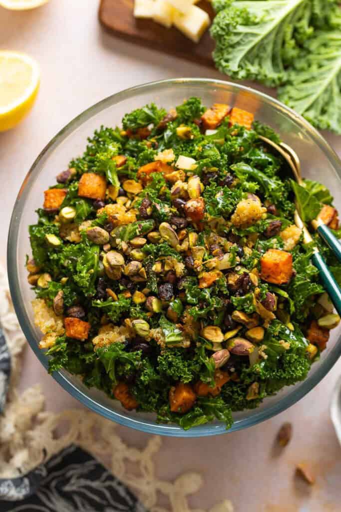 A salad made with roasted sweet potato & black beans, crispy quinoa, kale, cheese and chopped pistachios in a glass salad bowl with serving spoons.