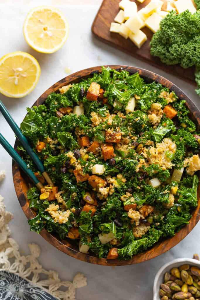 A salad made with roasted sweet potato & black beans, crispy quinoa, kale, cheese and chopped pistachios in a wooden salad bowl with serving spoons. Lemon halves, chopped cheese, raw kale and pistachios on the sides.