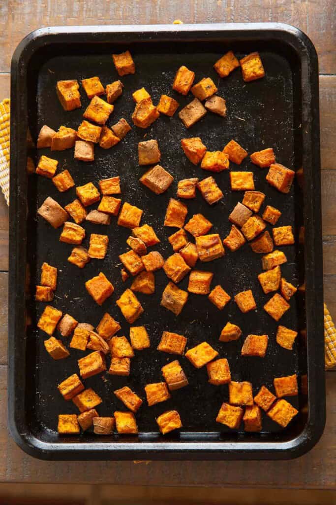 Roasted sweet potato cubes season in oil and spice, spread out on a backing sheet.