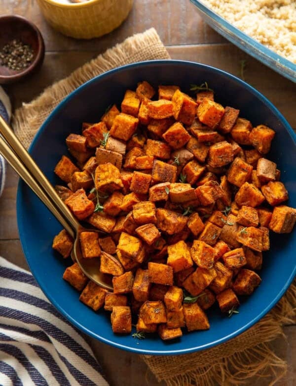 A serving dish with roasted sweet potatoes with serving spoons in the bowl and cooked quinoa on the side.
