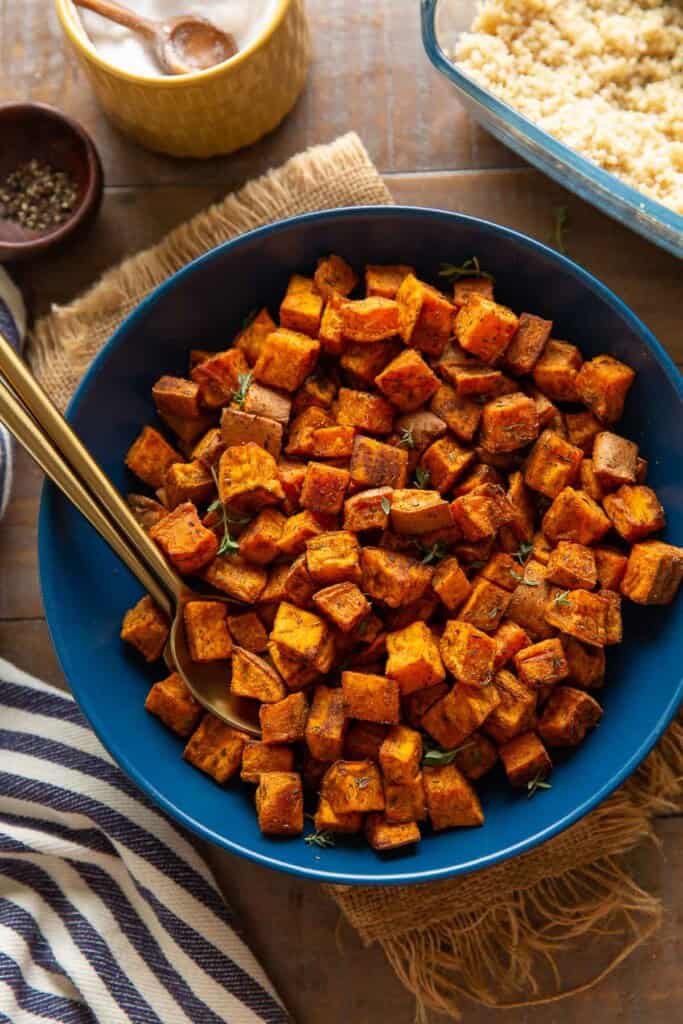 A serving dish with roasted sweet potatoes with serving spoons in the bowl and cooked quinoa on the side.