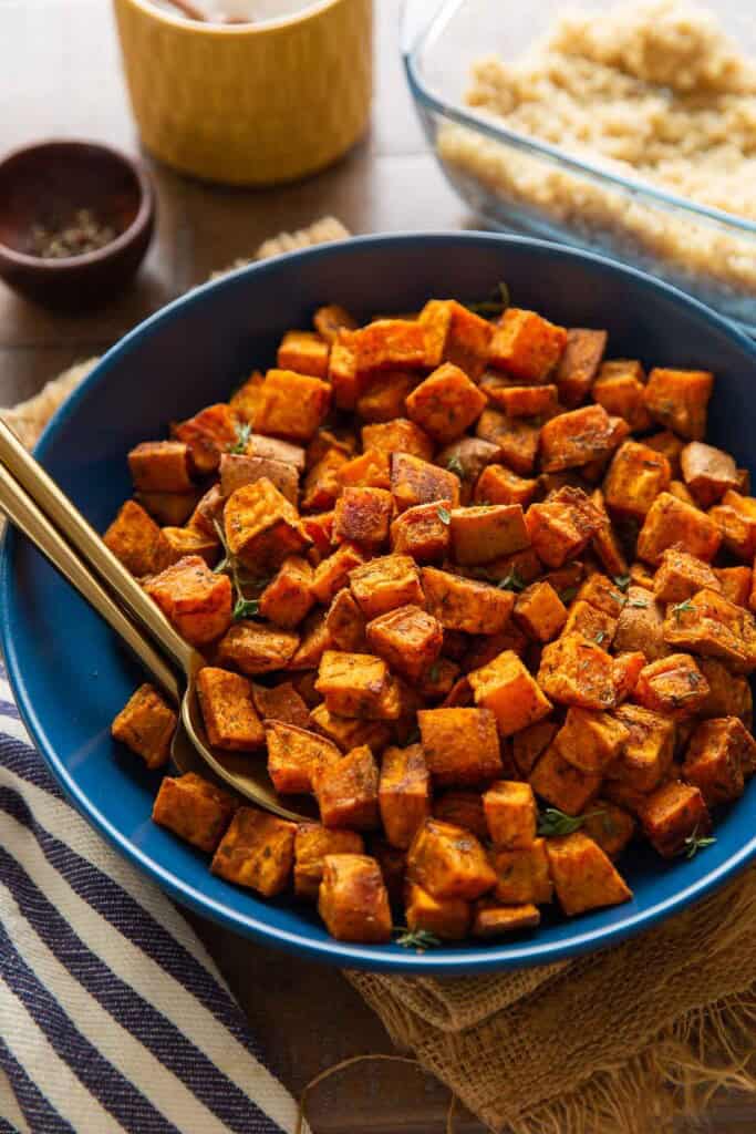 A serving dish full of roasted sweet potatoes with serving spoons in the bowl. Cooked quiona, salt and pepper on the side.