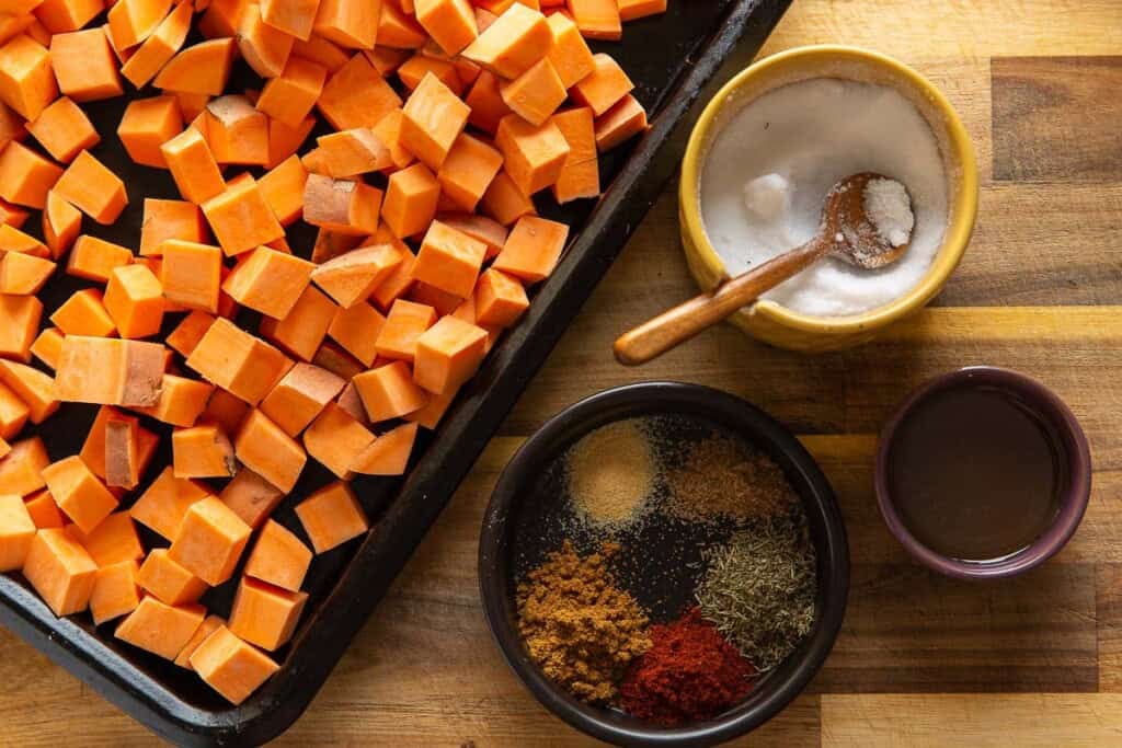 Diced, raw sweet potato on a baking sheet next to a salt bowl, oil and spices (garlic powder, cumin, coriander, chili powder and thyme).