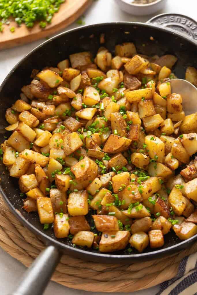 A cast iron skillet full of pan fried breakfast potatoes topped with fresh chopped chives and black pepper.