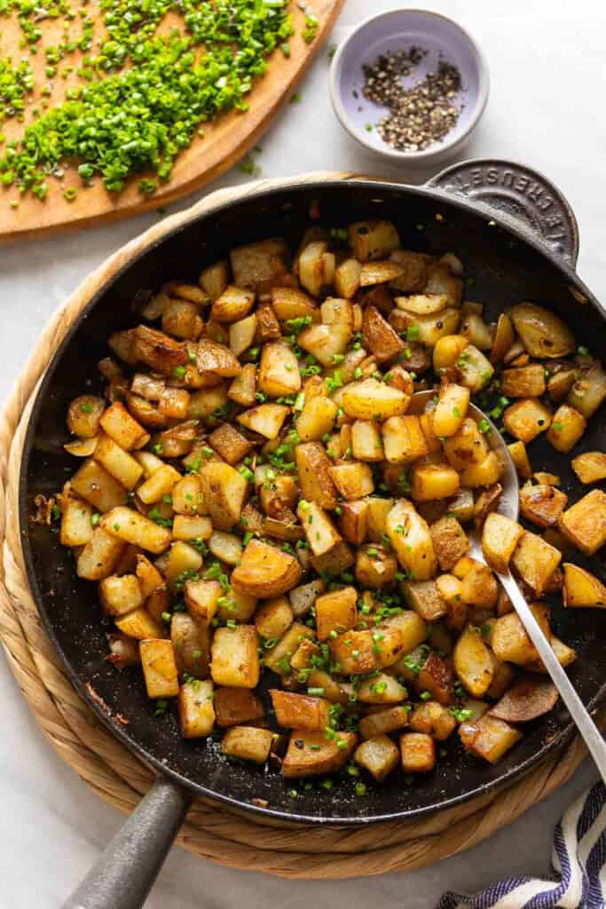Skillet Breakfast Potatoes topped with fresh chopped chives in a cast iron pan with a spoon. Fresh chopped chives and a small bowl of black pepper on the side.