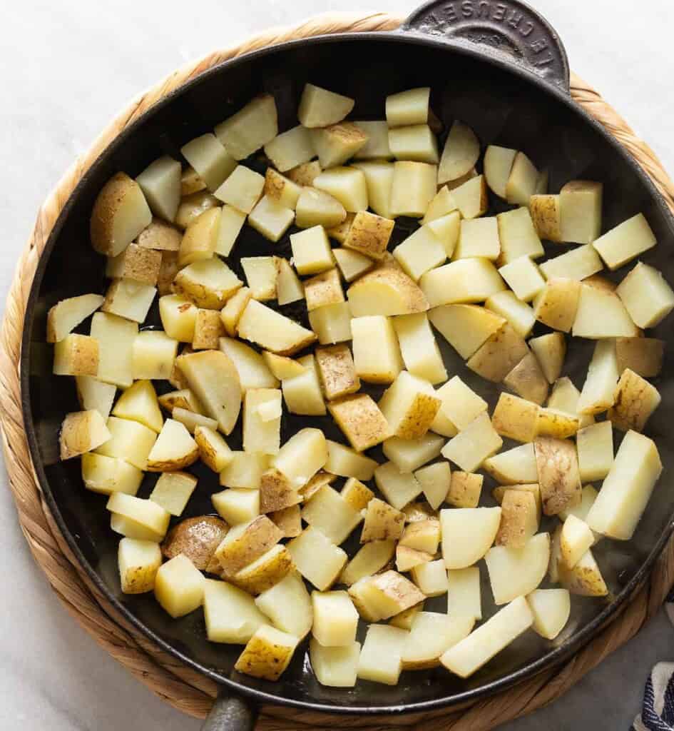 A large cast iron skillet with a single layer of par-boiled potatoes frying, untouched in oil to crisp up the bottom.