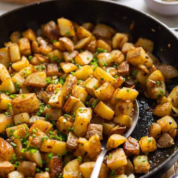 A spoon scooping up breakfast skillet potatoes topped with chopped chives in a cast iron pan.