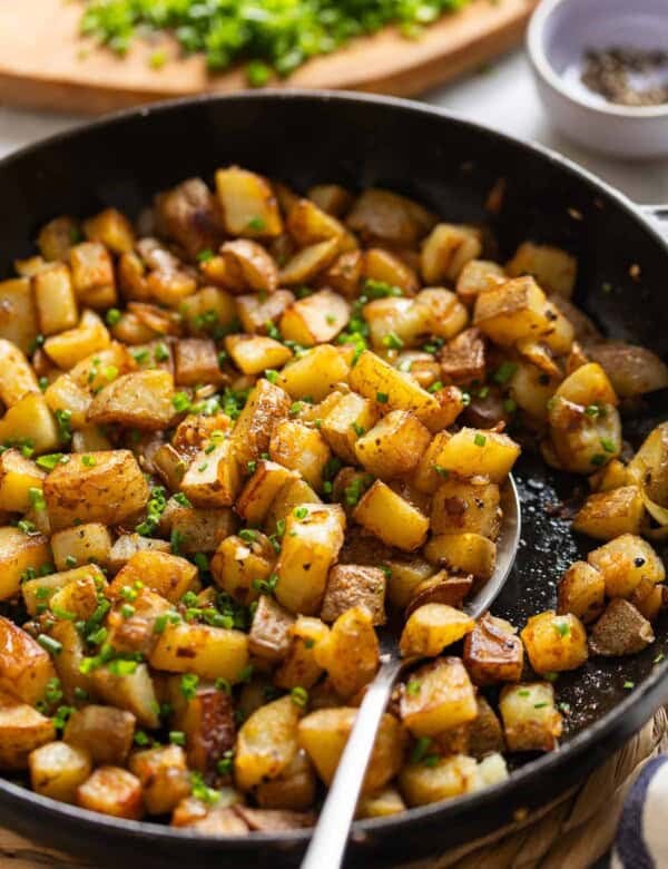 A spoon scooping up breakfast skillet potatoes topped with chopped chives in a cast iron pan.