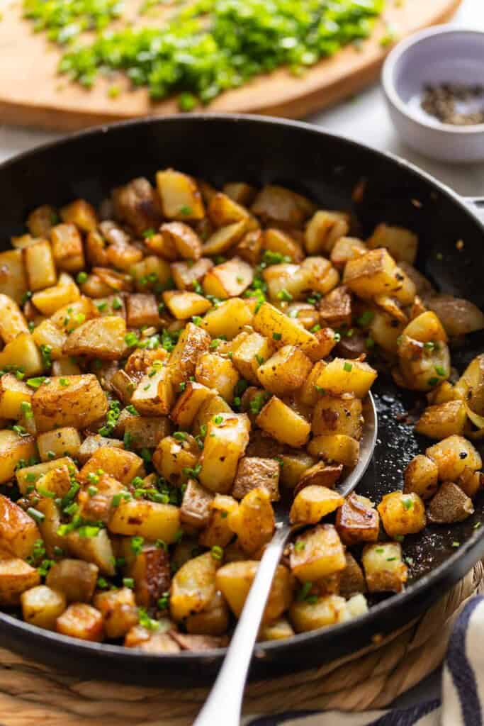 A spoon scooping up breakfast skillet potatoes topped with chopped chives in a cast iron pan.