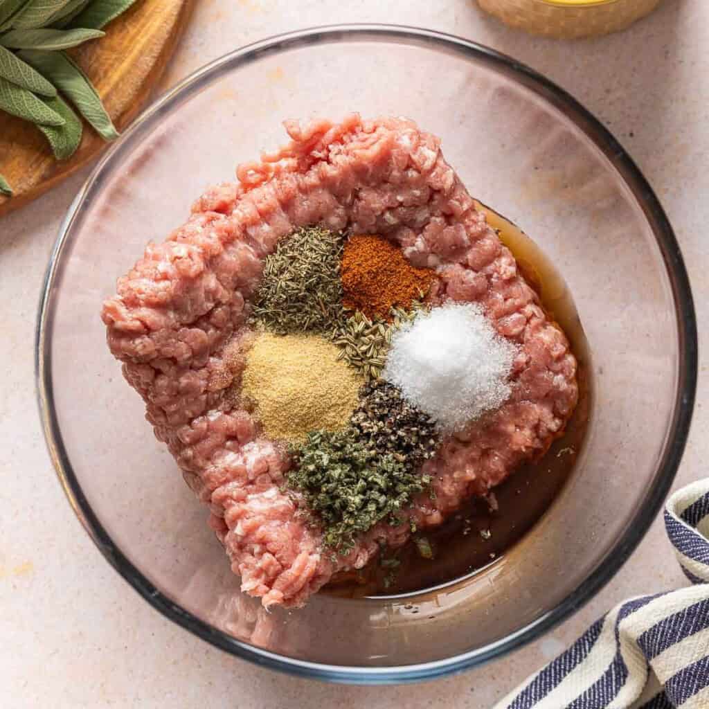 Turkey Maple Breakfast Sausage Patties ingredients in a mixing bowl: turkey mince, garlic powder, thyme, sage, salt, pepper, cayenne and maple syrup.