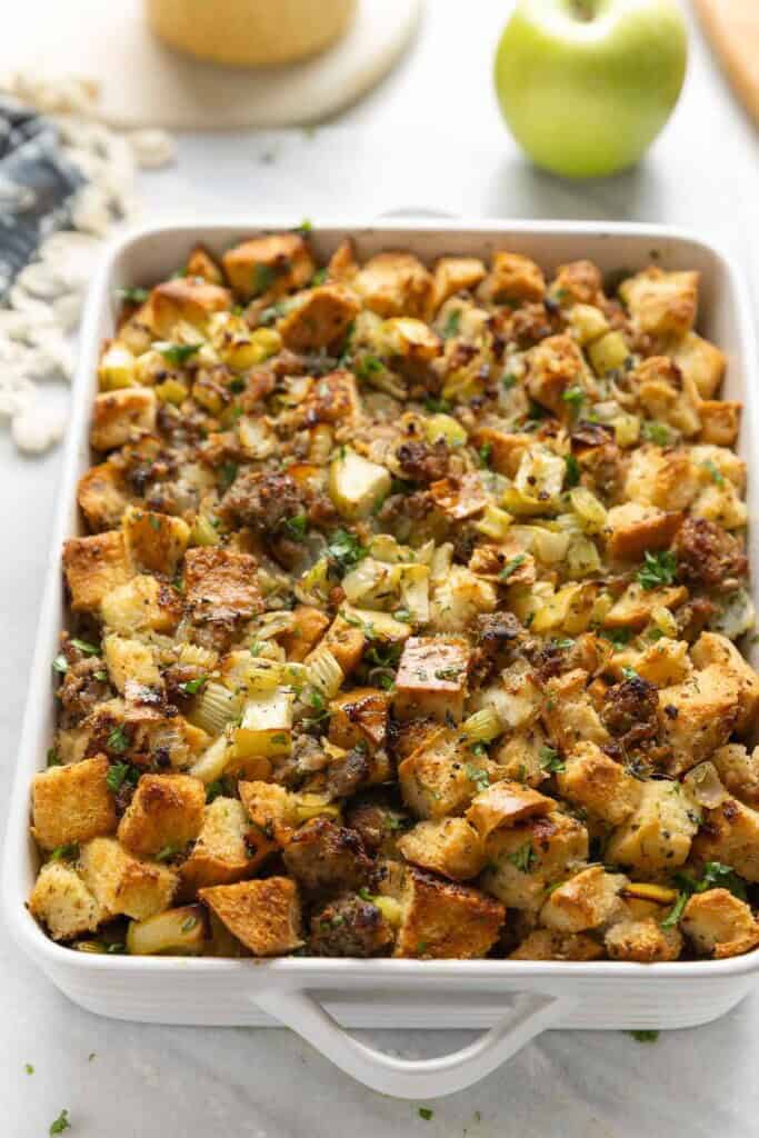 Gluten Free Stuffing with Sausage & Apple in a casserole dish with fresh chopped parsley on top with a kitchen cloth and apple in the back.