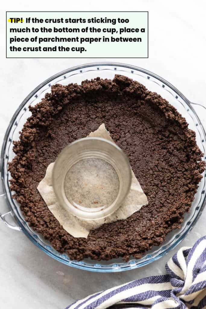 Gluten free oreo cookie crumbs pressing into a glass pie dish. "Tip! TIP! If the crust starts sticking too much to the bottom of the cup, place a piece of parchment paper in between the crust and the cup."