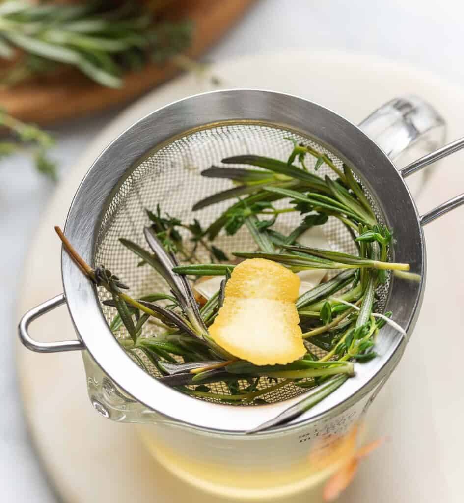 A fine mesh strainer straining out fresh herbs, citrus peel and a ginger tea bag.