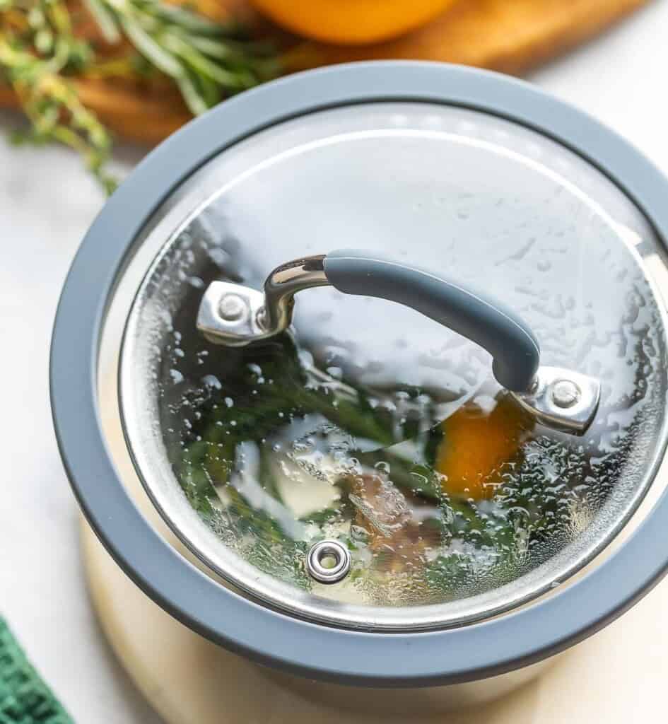 A lid covering a pot of water simmering with fresh herbs and orange peel