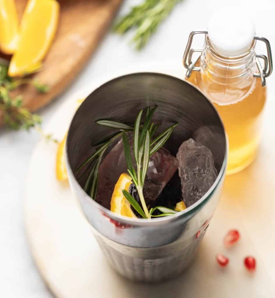 A cocktail shaker full of ice, fresh rosemary, thyme and orange peel