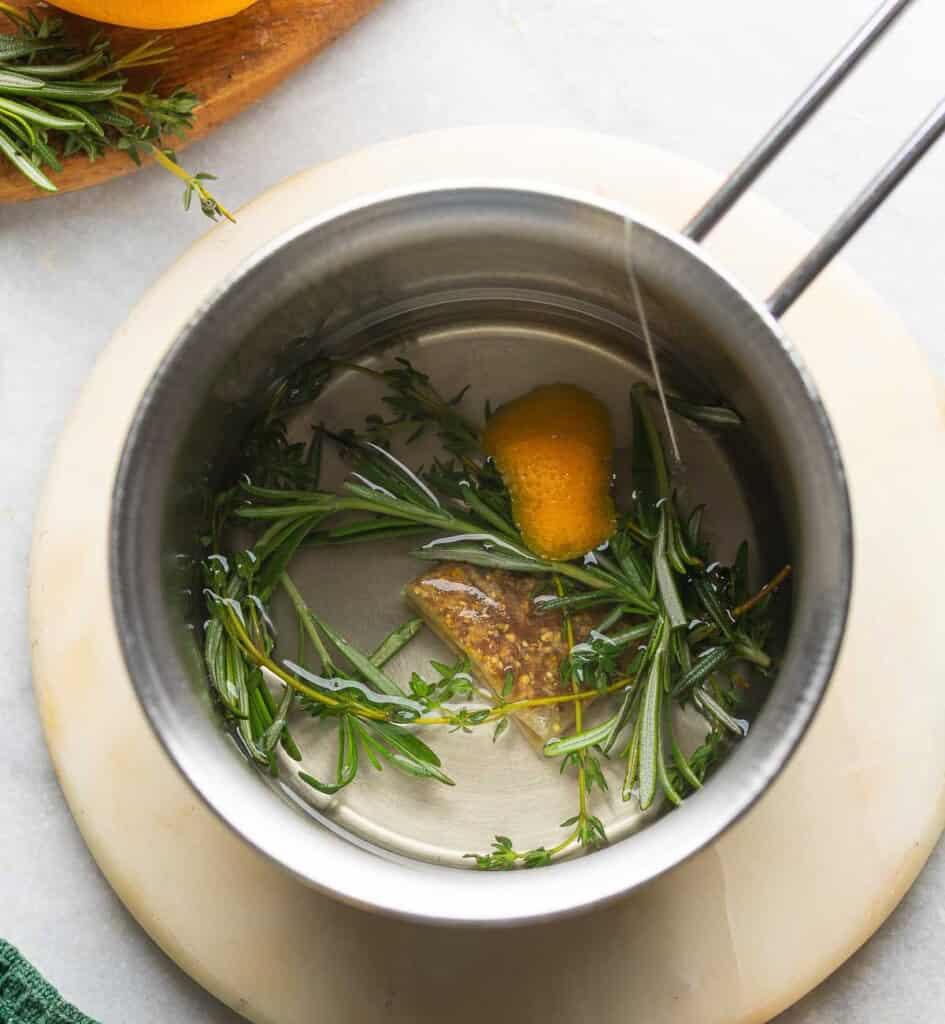 Hot water in a small pot with fresh herbs, orange peel and a ginger tea bag.