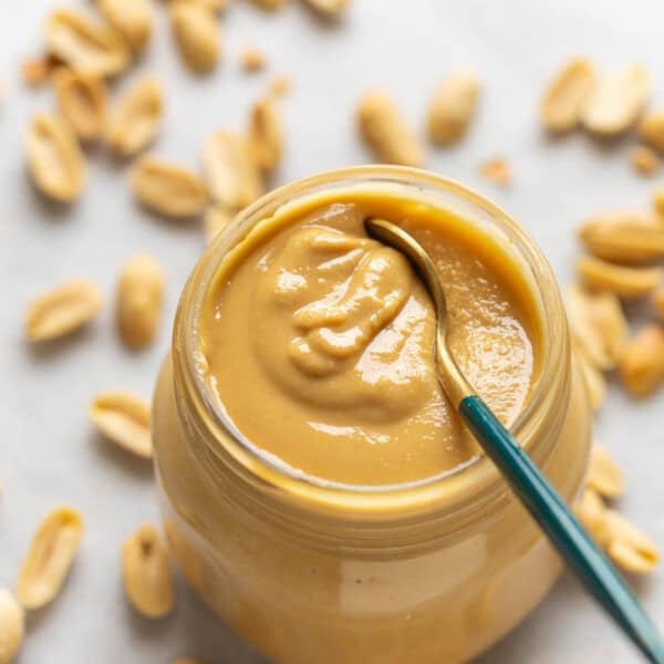 A spoon inside a jar of homemade peanut butter with roasted peanuts in the back