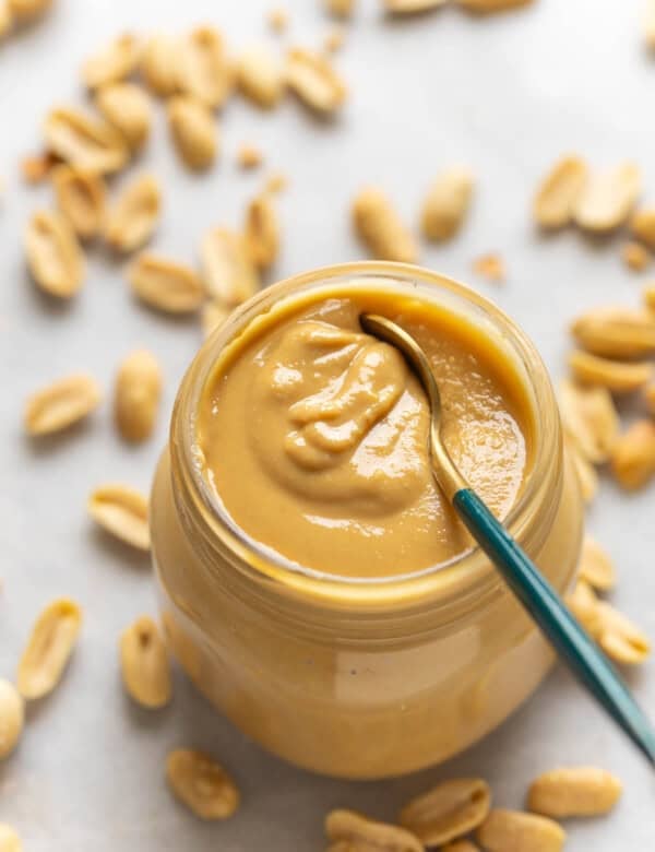 A spoon inside a jar of homemade peanut butter with roasted peanuts in the back