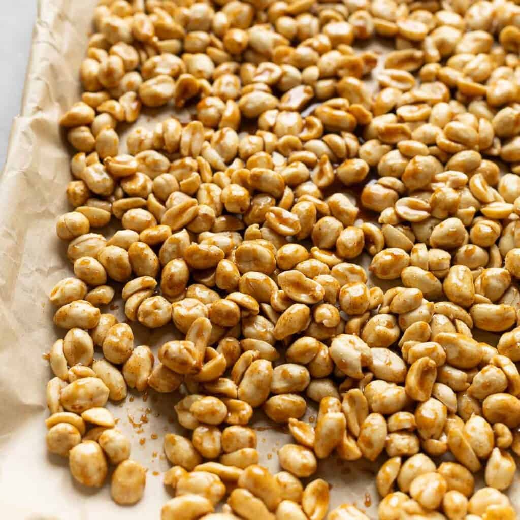 Lightly roasted peanuts coated in a sticky glaze spread across a parchment paper lined baking sheet.
