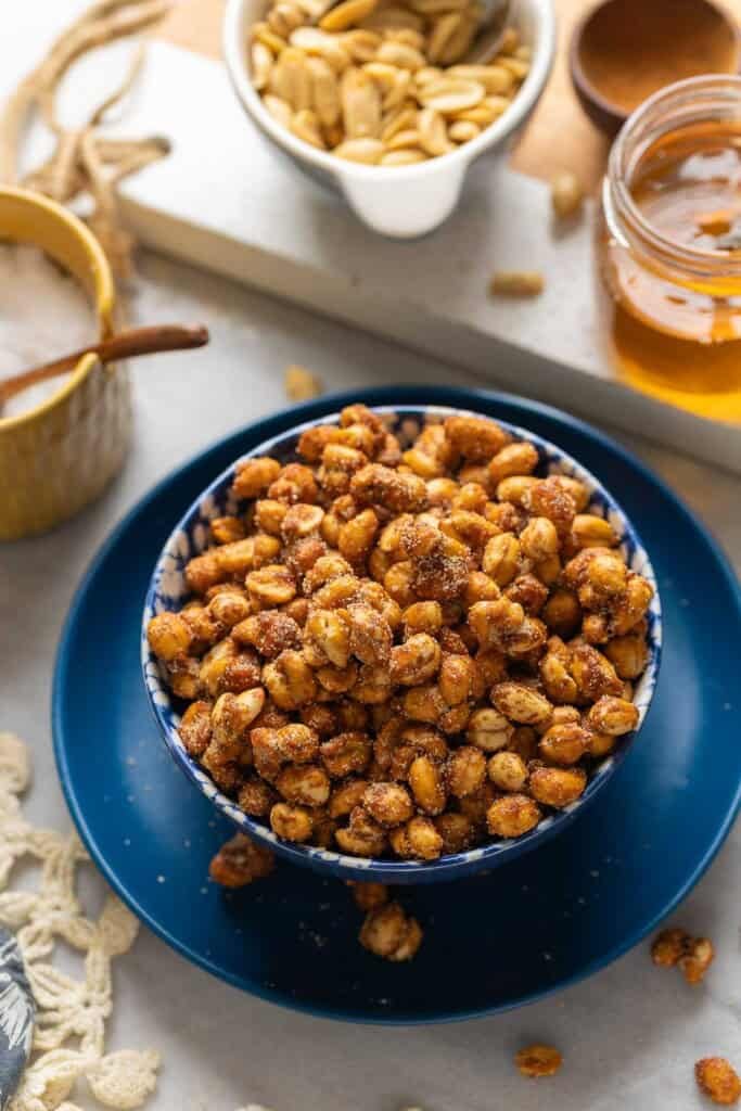 Honey Roasted Peanuts in a serving bowl with some of the whole ingredients in the back: salt, peanuts, cinnamon and honey