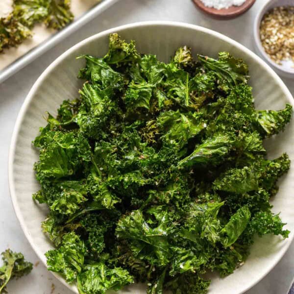 Homemade baked kale chips in a serving bowl with salt and za'atar spice in small bowls on the side and more kale chips in a baking sheet in the back