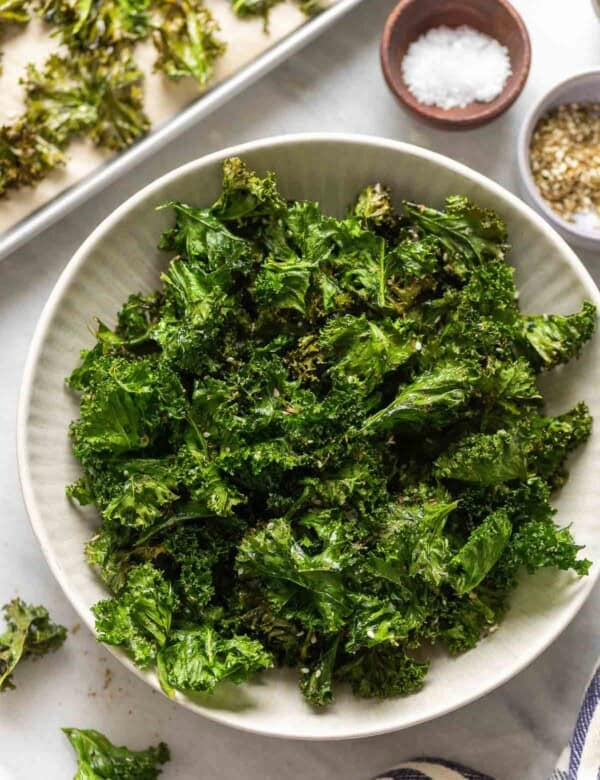 Homemade baked kale chips in a serving bowl with salt and za'atar spice in small bowls on the side and more kale chips in a baking sheet in the back