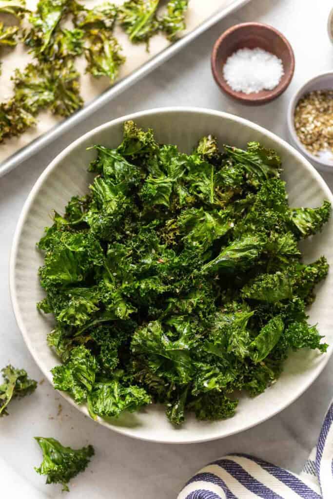 Homemade baked kale chips in a serving bowl with salt and za'atar spice in small bowls on the side and more kale chips in a baking sheet in the back