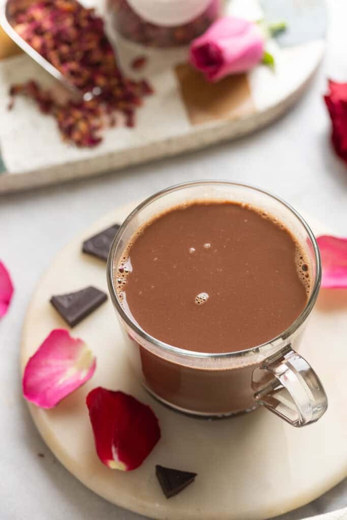A mug of rose hot chocolate surrounded by pink and red rose petals, dark chocolate pieces and rose leaf