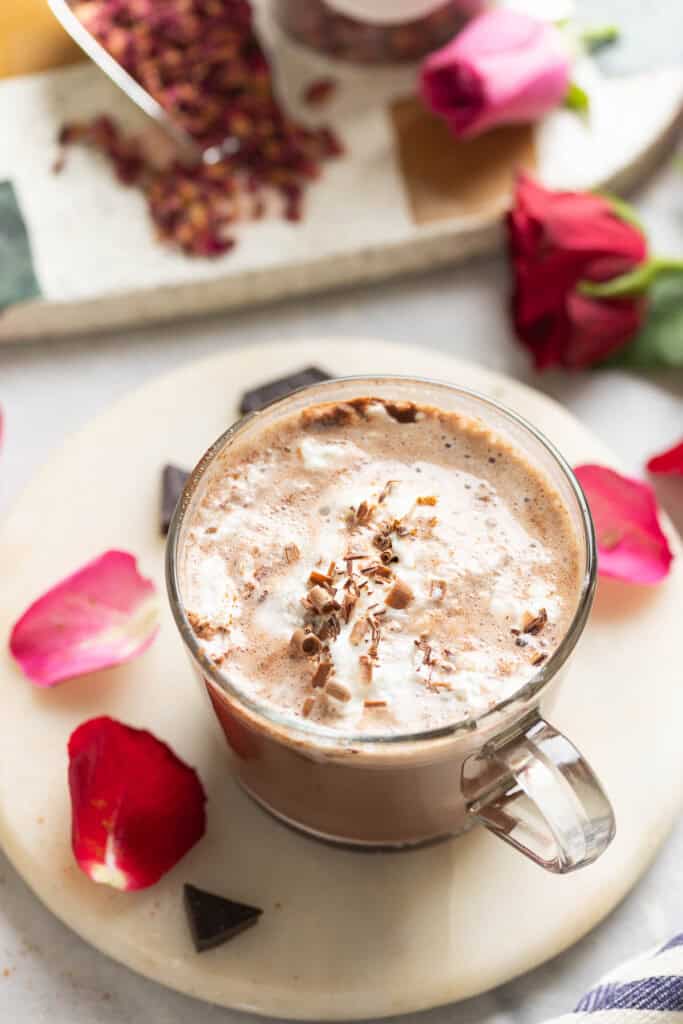 A hand grabbing the handle of a mug of rose hot chocolate topped with partially melted whipped cream and chocolate curls and surrounded by pink and red rose petals, dark chocolate pieces and rose leaf