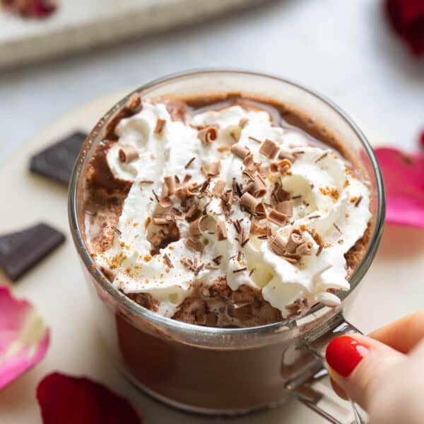 A hand grabbing the handle of a mug of rose hot chocolate with whipped cream and chocolate curls on top.