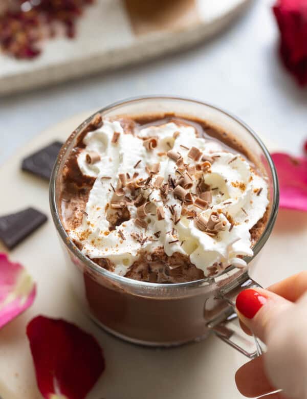 A hand grabbing the handle of a mug of rose hot chocolate with whipped cream and chocolate curls on top.