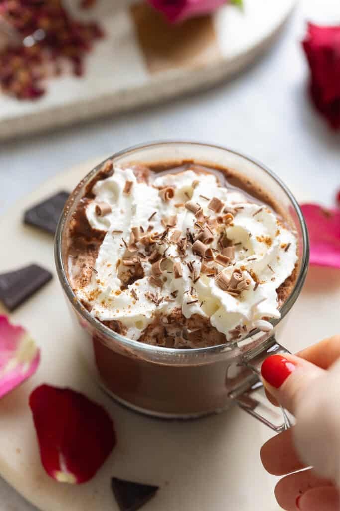A hand grabbing the handle of a mug of rose hot chocolate with whipped cream and chocolate curls on top.