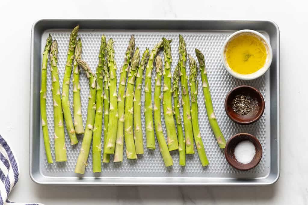 Raw asparagus on a baking sheet with bowls of salt, pepper and oil