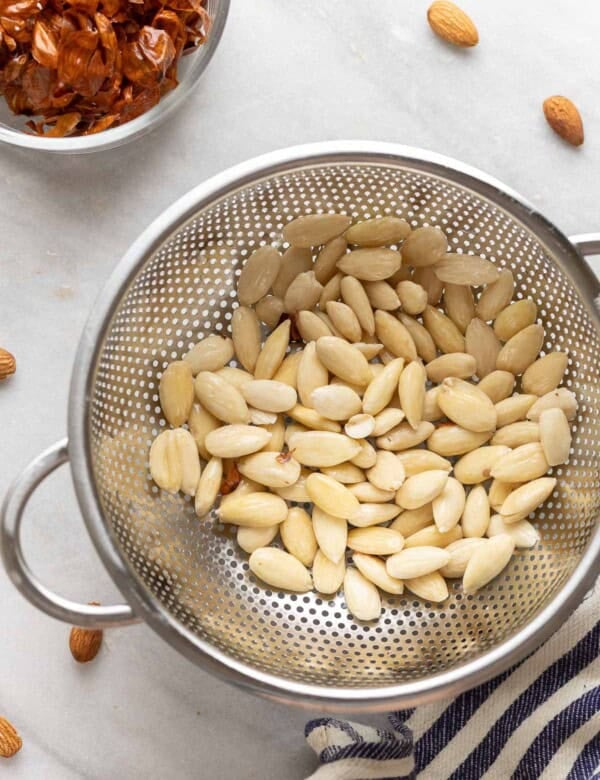 A colander full of blanched almonds with the almond skins in a separate bowl on the side.