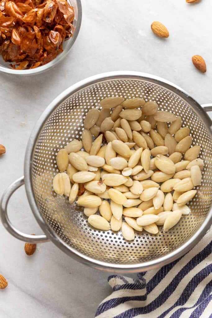 A colander full of blanched almonds with the almond skins in a separate bowl on the side.