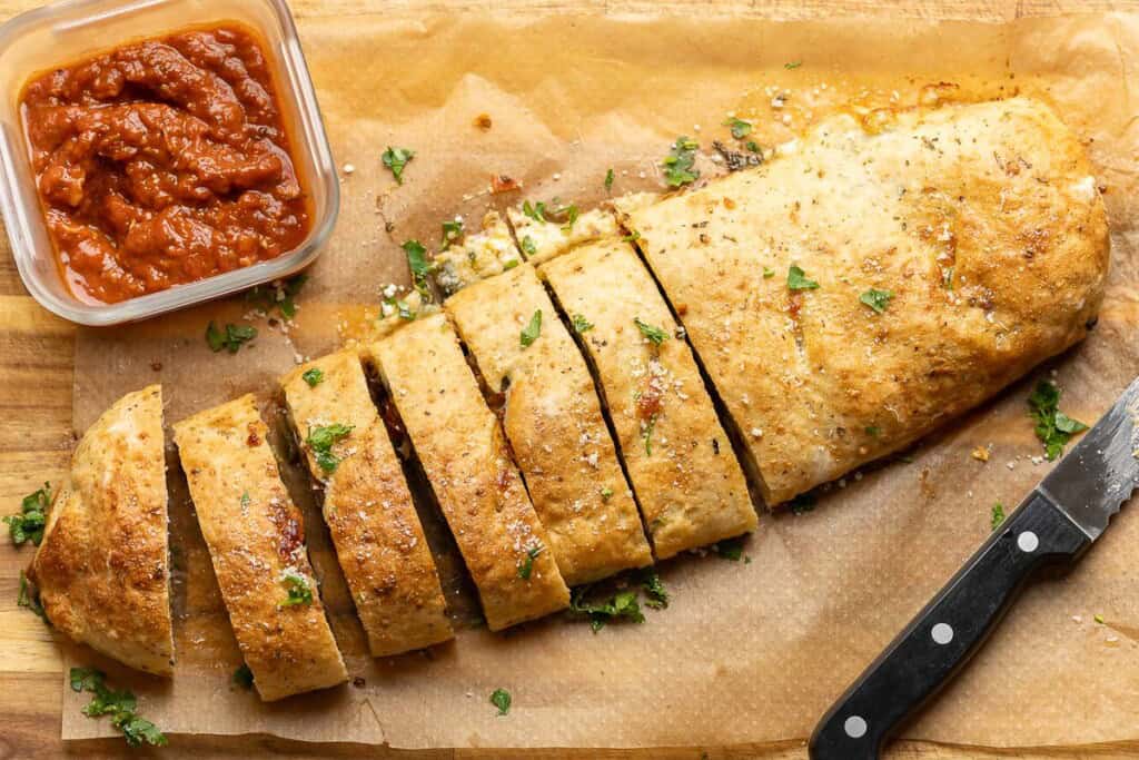 A Gluten free Stromboli log on a. wooden cutting board cut into slices with a small container or marinara on the side.