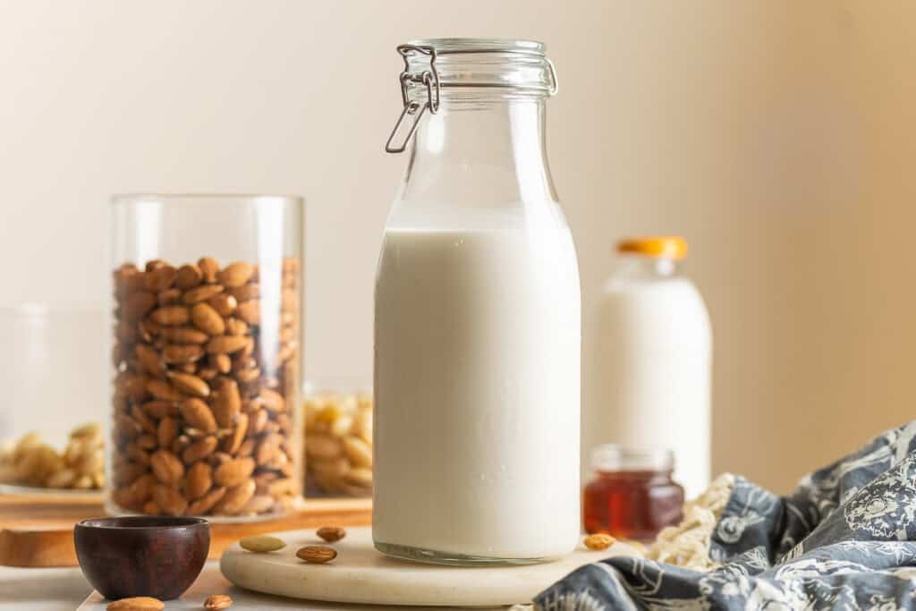 Homemade almond milk a large flip top jar with raw blanched and un blanched almonds in other jars. A small glass of maple syrup an a milk bottle in the back.