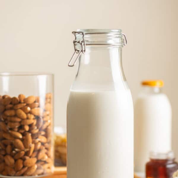 Homemade almond milk a large flip top jar. Raw almonds in another jar and scattered around the milk. A small glass of maple syrup an a milk bottle in the back.