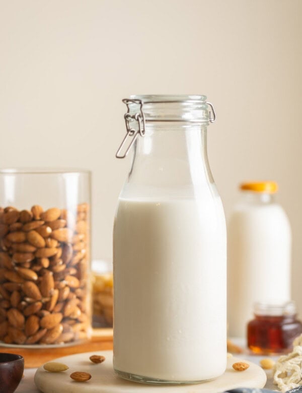 Homemade almond milk a large flip top jar. Raw almonds in another jar and scattered around the milk. A small glass of maple syrup an a milk bottle in the back.