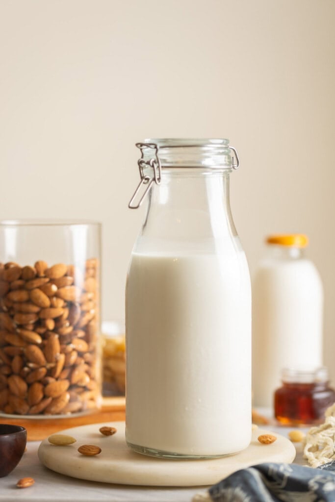 Homemade almond milk a large flip top jar. Raw almonds in another jar and scattered around the milk. A small glass of maple syrup an a milk bottle in the back.