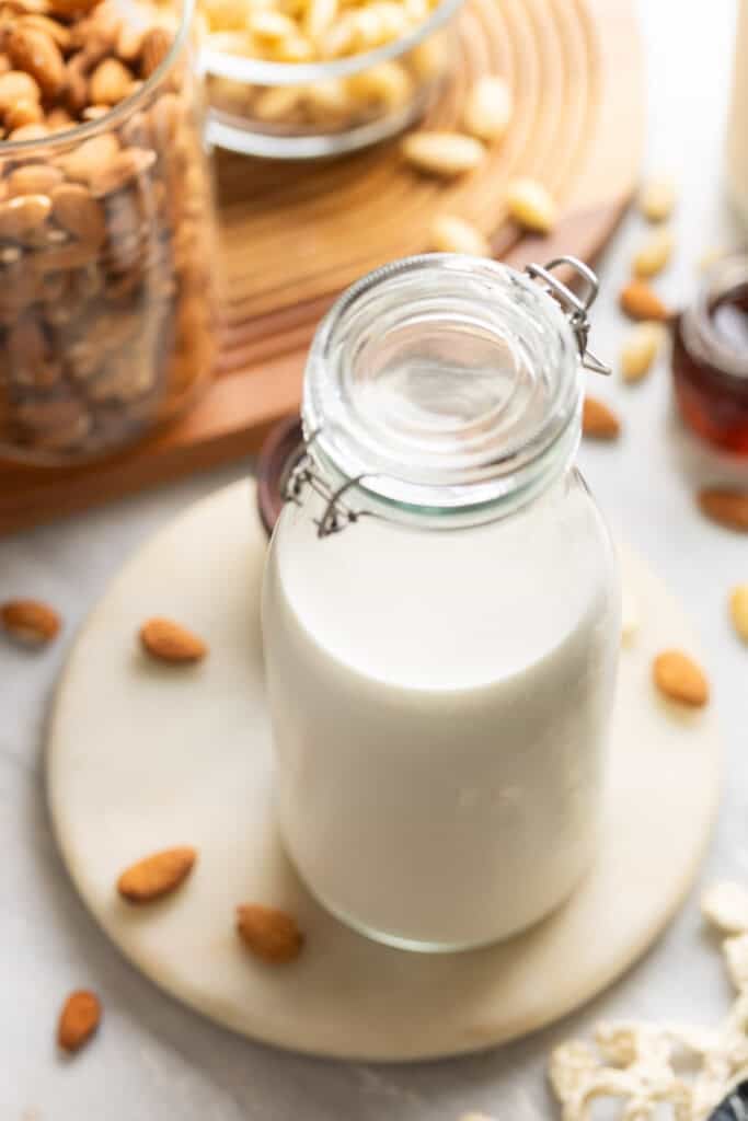 A large glass jar full of homemade almond milk with scattered almonds all around.