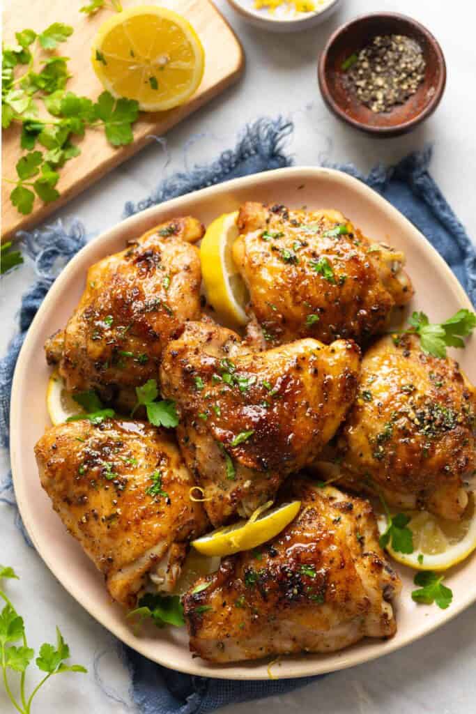 Lemon Pepper Chicken Thighs on a serving plate with lemon slices and fresh parsley and small bowl of black pepper on the side.