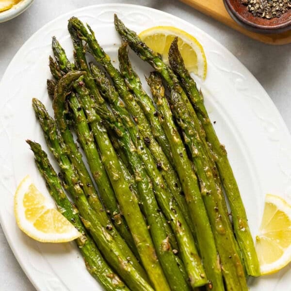 Oven Roasted Asparagus on a serving plate with lemon slices tucked along side the roasted stalks and a small bowl of black pepper on the side