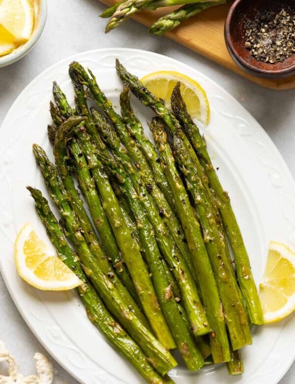 Oven Roasted Asparagus on a serving plate with lemon slices tucked along side the roasted stalks and a small bowl of black pepper on the side