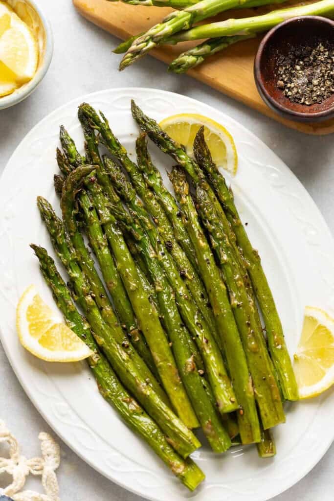 Oven Roasted Asparagus on a serving plate with lemon slices tucked along side the roasted stalks and a small bowl of black pepper on the side
