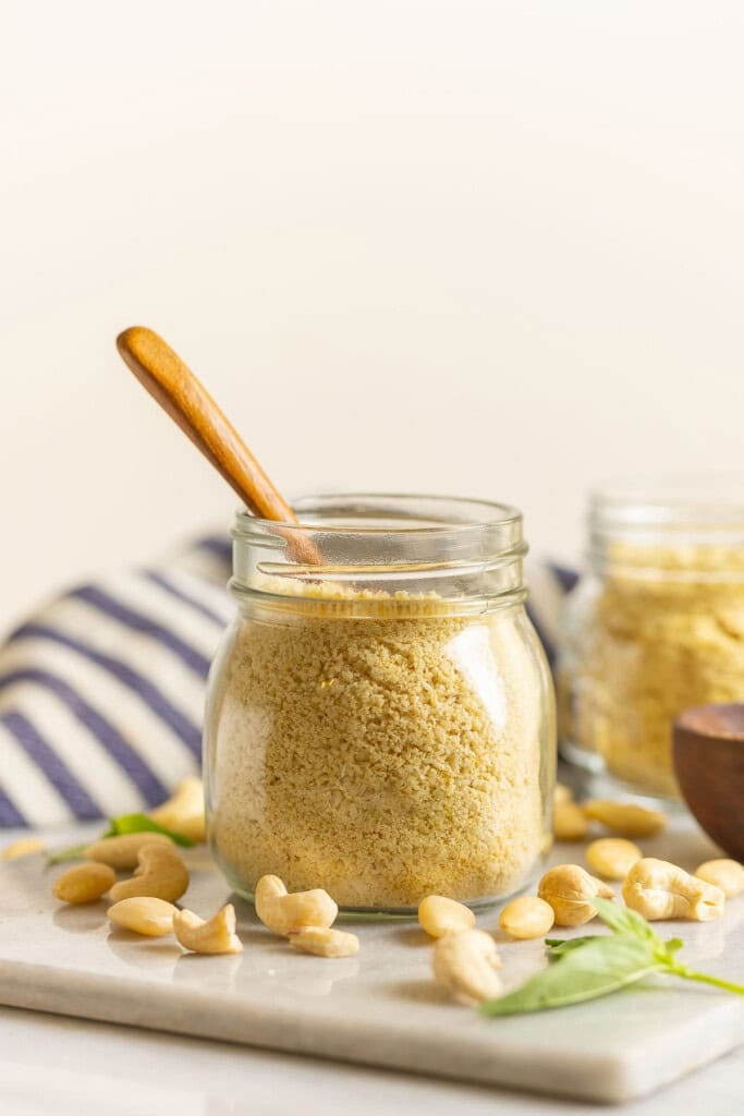 Homemade vegan parmesan cheese in a jar with a wooden spoon inside and surrounded by raw almonds and cashews.
