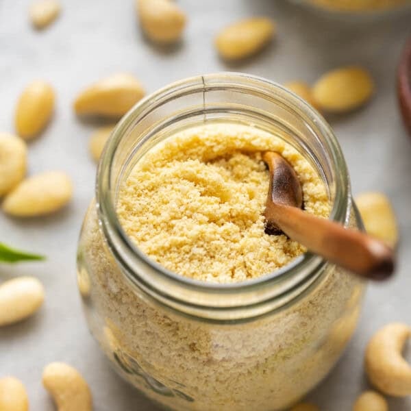 A small jar of Homemade vegan parmesan cheese with a little wooden spoon inside surrounded by cashews and almonds