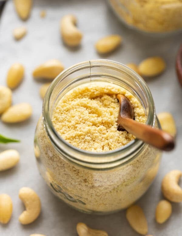 A small jar of Homemade vegan parmesan cheese with a little wooden spoon inside surrounded by cashews and almonds